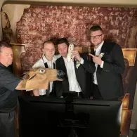 Four men in tuxedos pose behind a monitor, one wearing a horse head mask, at a formal event