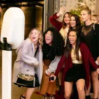 Five women posing and smiling outdoors at night, one with her hand raised in excitement