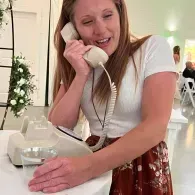 Woman in a white top holding a white phone handset at a counter, smiling in a bright room.