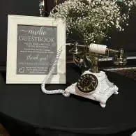 Vintage guestbook table with framed sign, white rotary phone, and flowers on a black tablecloth