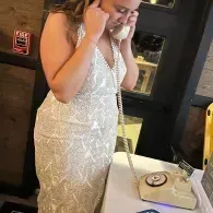 Woman in a white dress speaking on a phone beside a table with a beige handbag.
