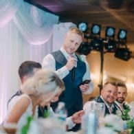 Wedding guest raising a glass at a reception table, with blurred guests and monitors in the background