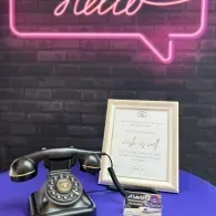 Vintage black rotary phone on a purple table beside a framed sign, with a pink neon 