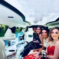 Four women in a white limousine, dressed in formal attire, smiling at the camera.