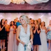 Bride in white gown holding bouquet at wedding reception with guests applauding in the background