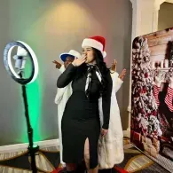 Woman in black dress and Santa hat posing at a holiday event beside a ring light and festive backdrop