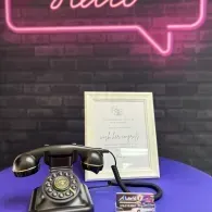 Vintage black rotary phone on purple table with framed certificate and neon sign against brick wall