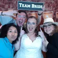 Four friends pose at a bridal party, with a “Team Bride” sign and a white wedding dress center stage.