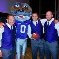 Four men in blue vests pose with a large cat mascot in a sports jersey holding a football.