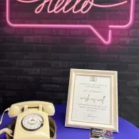 Cream rotary phone and framed certificate on a blue table beneath a pink neon “Hello” sign.