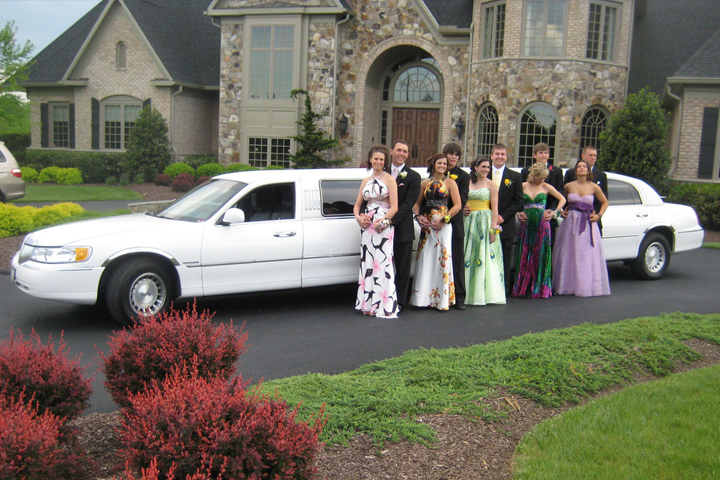 Group posing beside white limousine in front of a large house, wearing formal dresses.