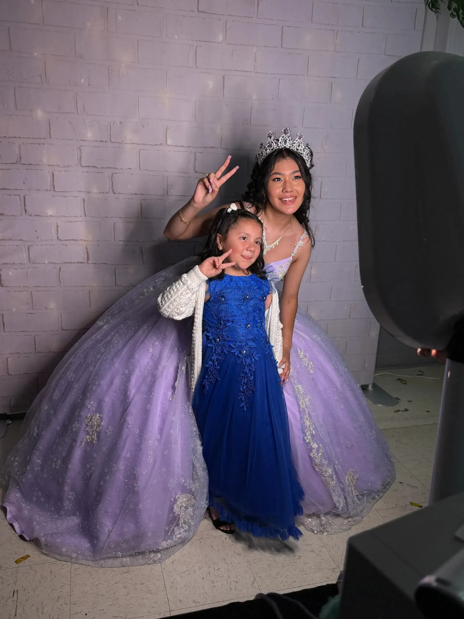 Person in a lavender gown poses with a child in a blue dress against a gray studio backdrop.