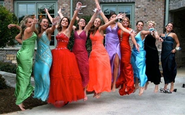 Group in colorful formal dresses jumping and posing outdoors in front of a building.