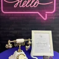 Vintage telephone and framed sign on a purple table beneath a pink neon “Hello” sign on a brick wall.