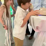Two children standing by a table at a party, speaking on a vintage rotary phone.