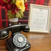 Vintage black rotary phone on wooden table beside framed certificate and yellow-red flowers