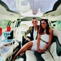 Two people sitting in a white limousine, smiling and wearing sunglasses