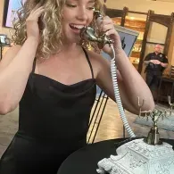 Woman in a black dress smiling while talking on a vintage phone at a table with a lace cloth