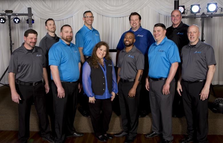 Group of nine people posing indoors in coordinated blue and gray shirts under stage lights.