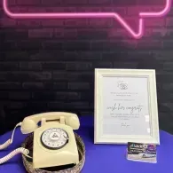 Vintage cream rotary phone, framed certificate, and small sign on a purple table beneath a pink neon light.