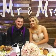 Couple seated at a decorated wedding reception table with “Mr & Mrs” sign behind them.