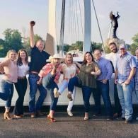 Group of people posing outdoors on a bridge, smiling with raised arms beside a tall statue.