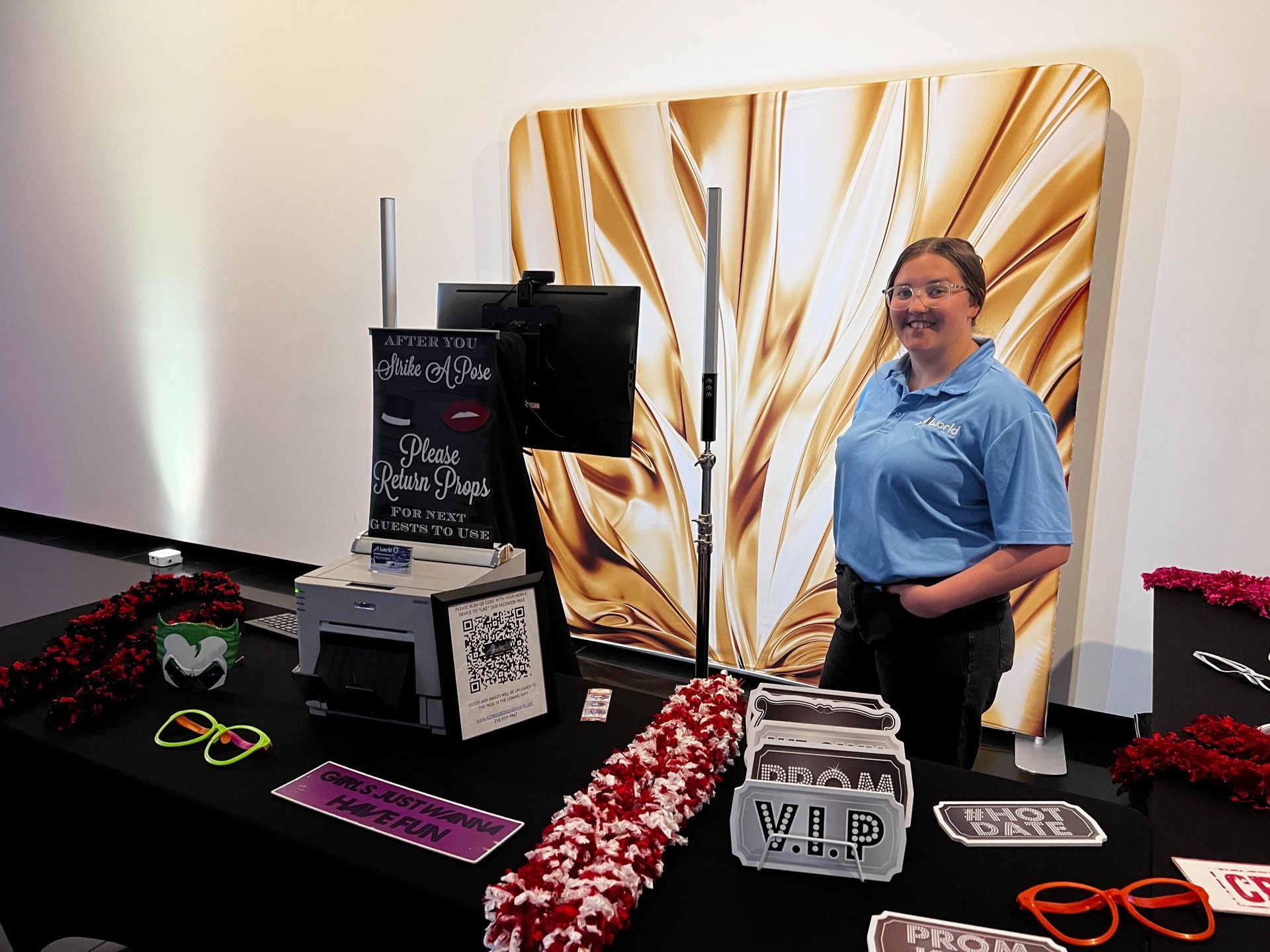 Photo prop booth with items displayed and a smiling woman beside a gold abstract backdrop.