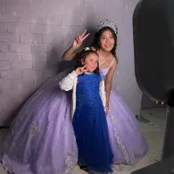 Two smiling people posing in lavender and blue formal dresses against a gray backdrop