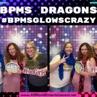 Three cheerleaders pose with pom-poms and signs in front of a purple lit backdrop, “BPMS DRAGONS” text above.