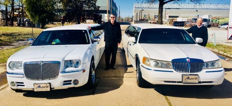 Two white luxury limousines parked side by side with two people standing between them outdoors