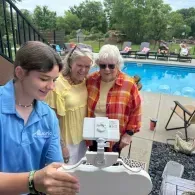 Three women by a pool looking at a white mobile device on a cart