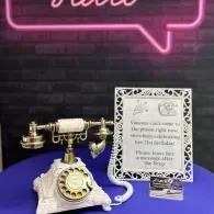 Vintage cream rotary phone beside a framed sign and tip jar on a purple table under neon lights