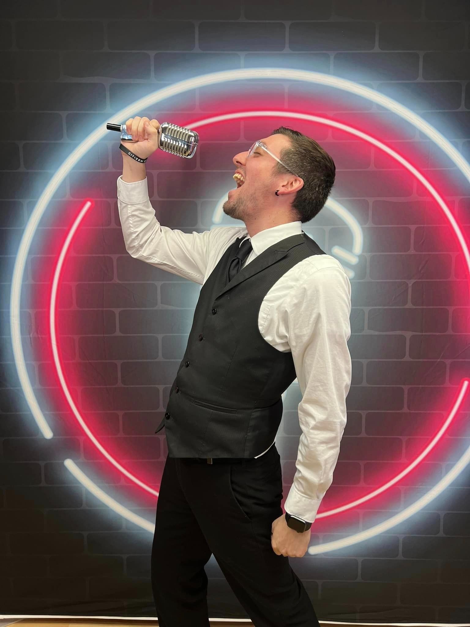 Man in vest and tie singing into a microphone before a glowing neon ring backdrop