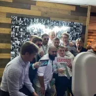 Group posing indoors in front of a black-and-white photo wall, holding signs and smiling.