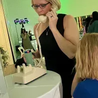 Woman in black dress using a cream telephone at a table with a mirror and flowers