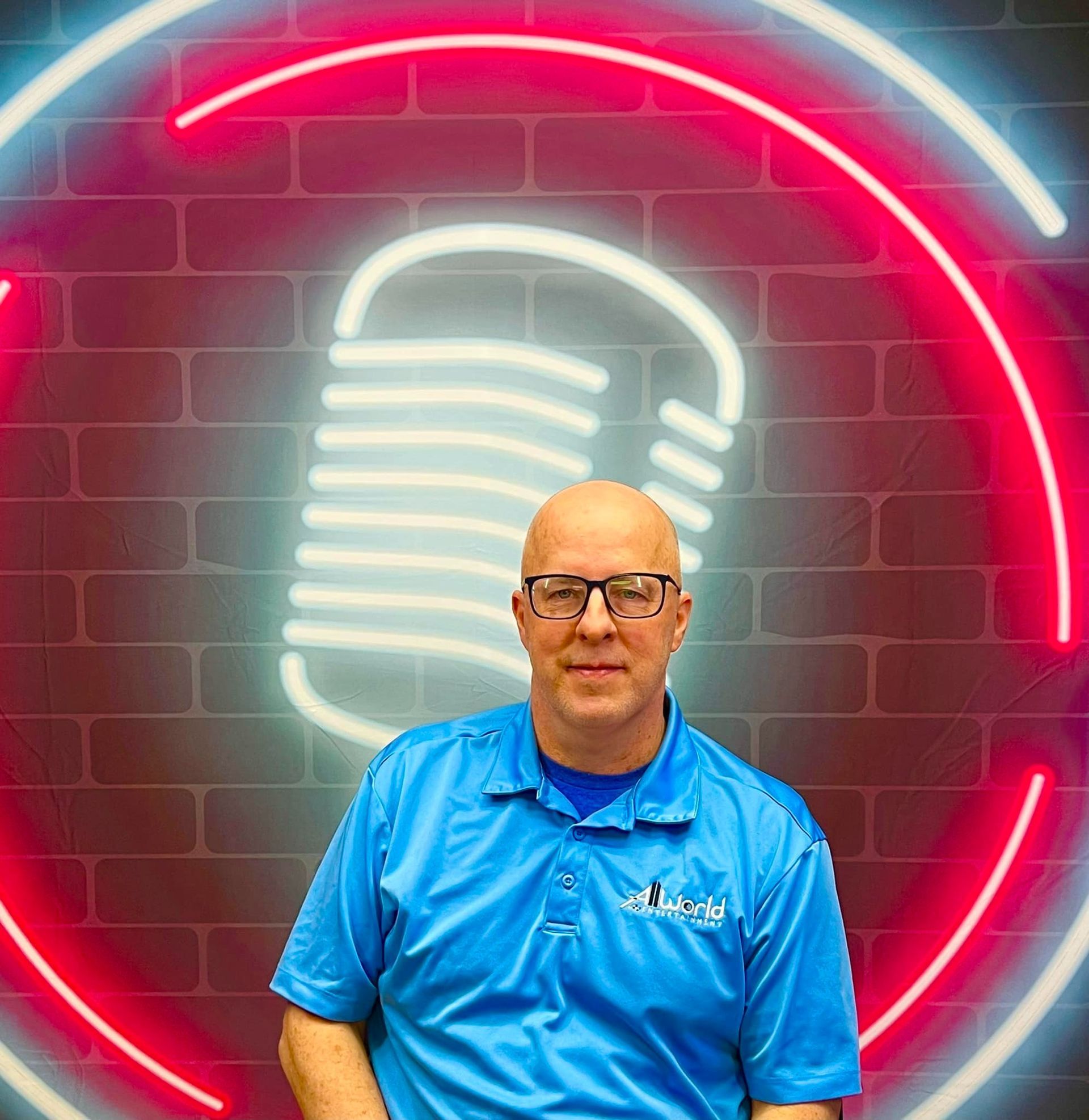Man in blue polo shirt standing before a neon microphone sign on a brick wall