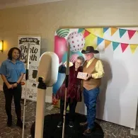 Two people posing in a photo booth with colorful bunting and a “Photo Booth” sign