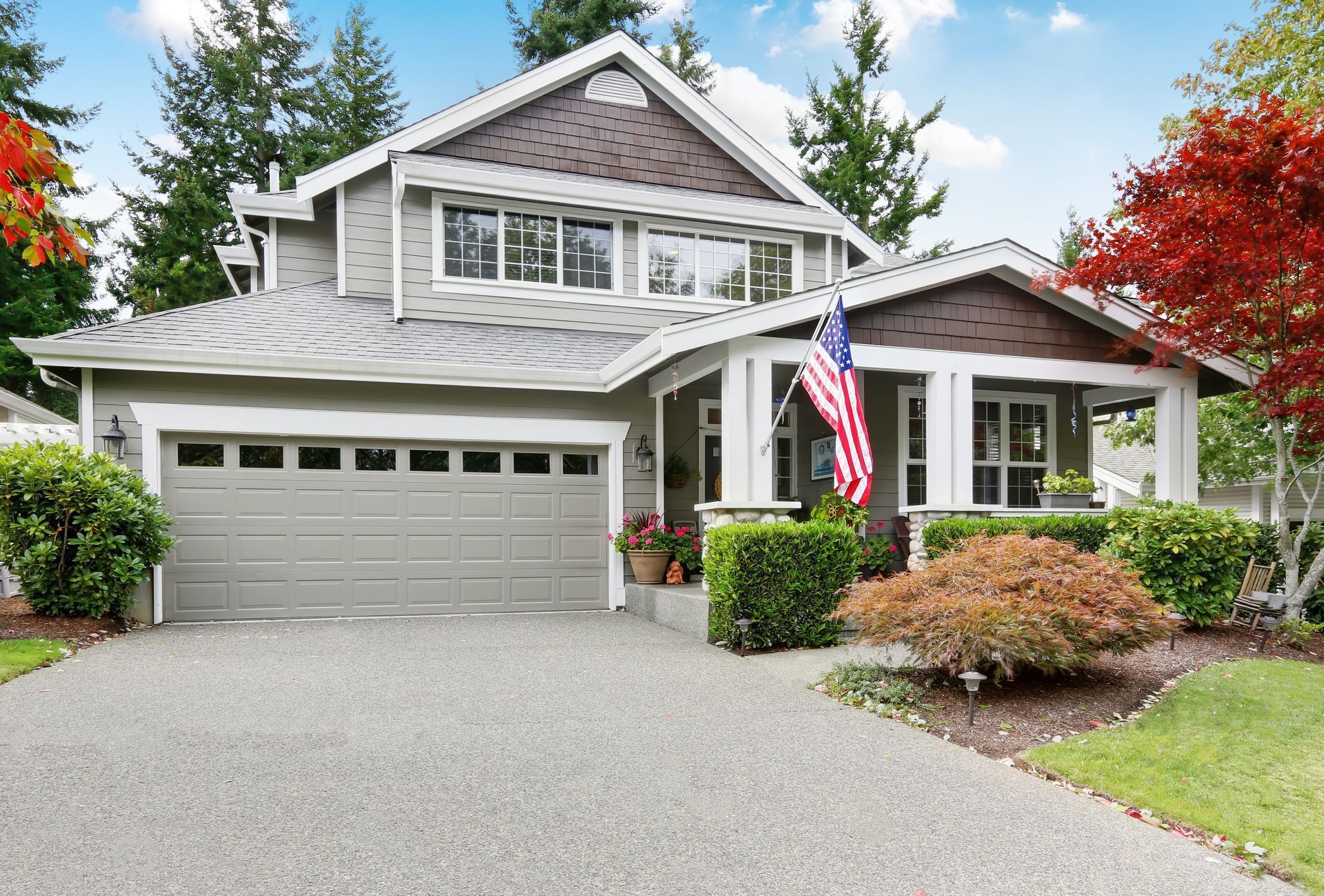 Suburban two-story gray house with attached garage, front porch, and American flag amid green landscaping