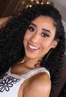 A woman with curly hair is wearing a white top and smiling.