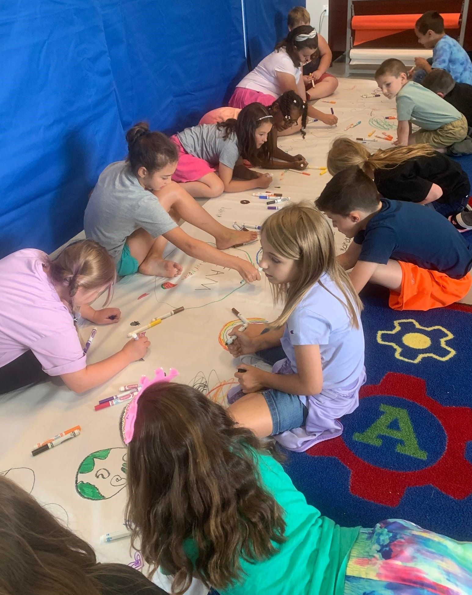 Club kids sitting on the floor in two lines drawing on a white canvas.