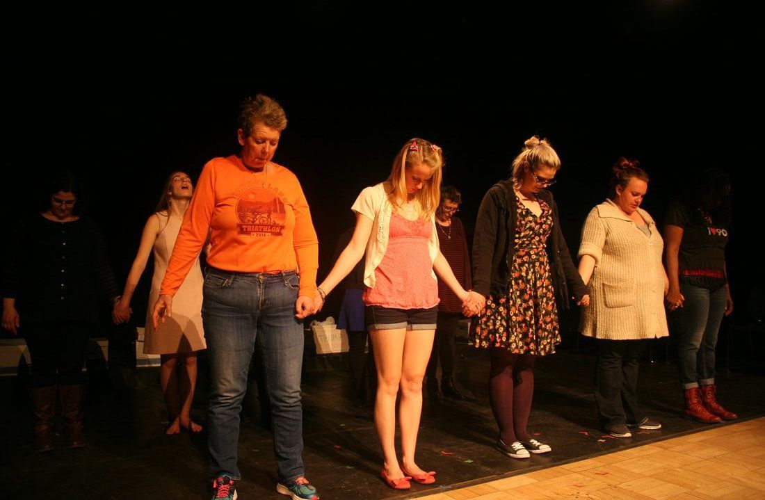 Community theater participants and Jayme Kilburn holding hands together on stage with their heads bowed.