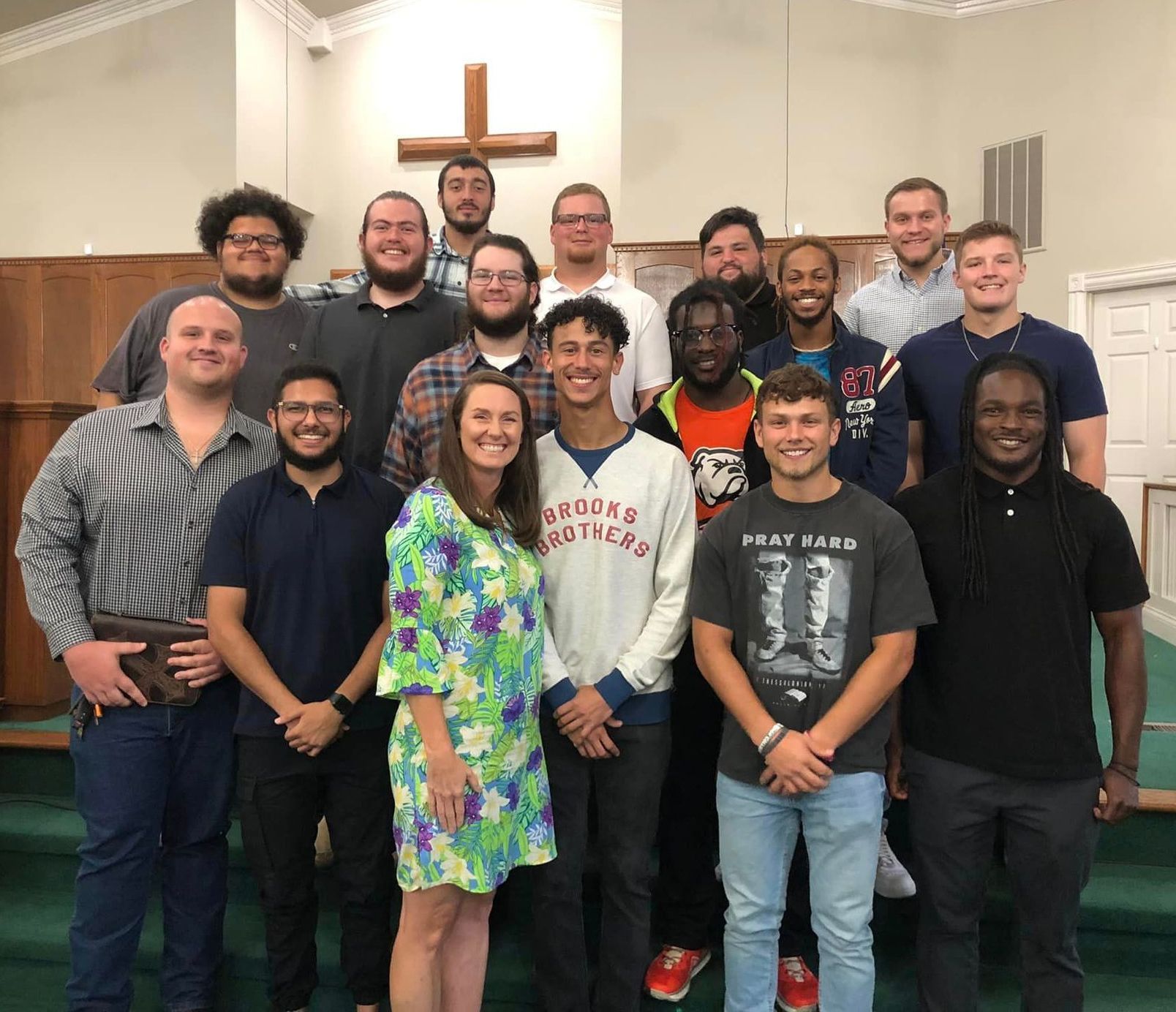 Tyce stands in the center of a group of Union students and staff smiling at the camera. They are standing in rows on green carpeted stairs, and a large wooden cross is visible on the wall behind them. 