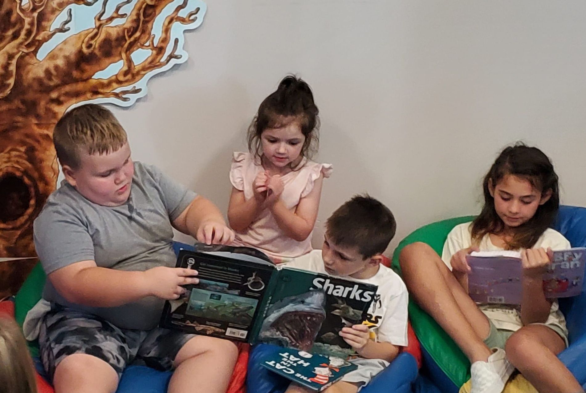 Four Club kids read books together, sitting on red, green, blue, and yellow bean bags with a drawing of a tree with a hole in the center visible behind them. Two kids hold a book with a large shark on the cover while a third looks on behind them. To their right, another child reads a book with a purple cover. 