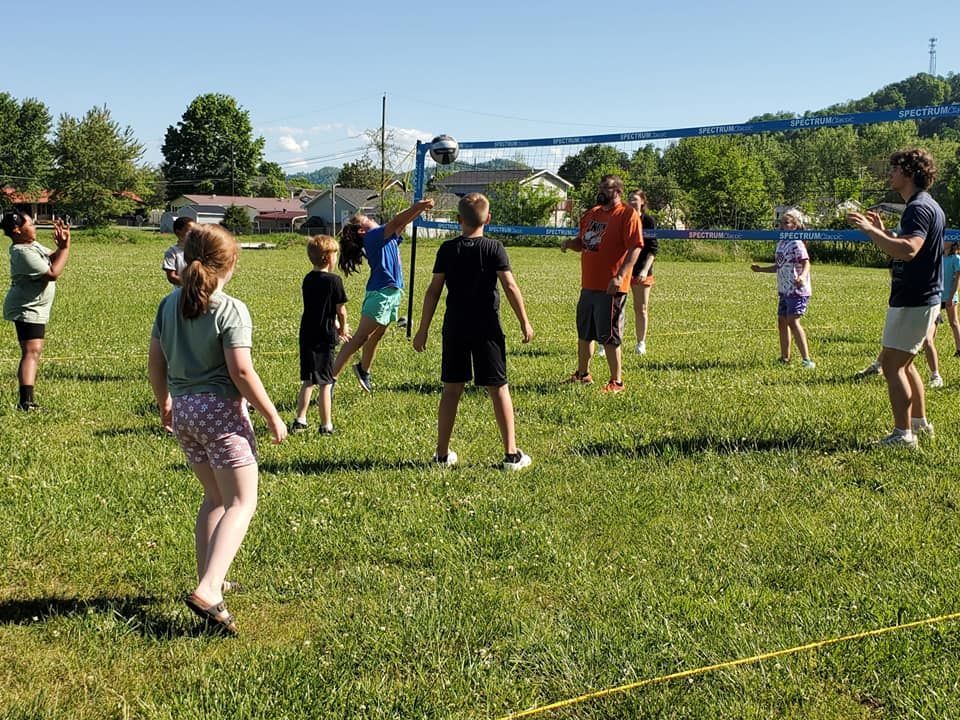 Union Club kids gather around a volleyball net in a field of bright green grass with Coach Jeremy Wise and Athletic Trainer Shea Hoffman. Everyone is wearing t-shirts and shorts, and one child is jumping to strike a descending volleyball. A few houses are visible in the background.