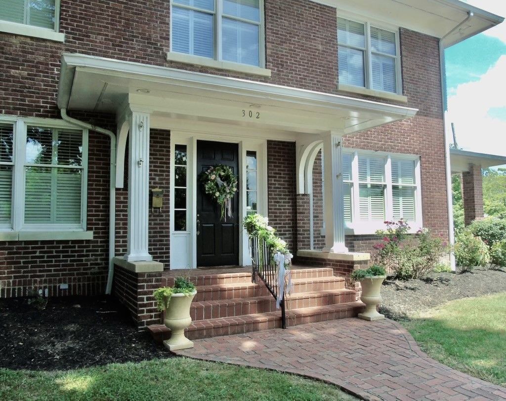 Front view of Baldwin Place, a brick home with white trim, brick front steps, and a brick front walk. The front door is decorated with a floral wreath, a floral garland lines the cast iron porch railing, and plants in urn-style flower pots are placed at the bottom of the steps.