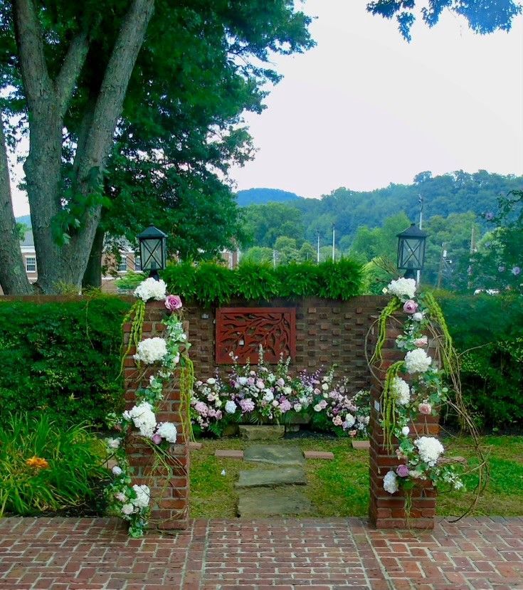 A scene from the wedding site: The brick-lined patio and pillars are adorned with white and pink floral decorations. Behind this is a brick wall with flowers in front of it and a view of the mountains in the background.