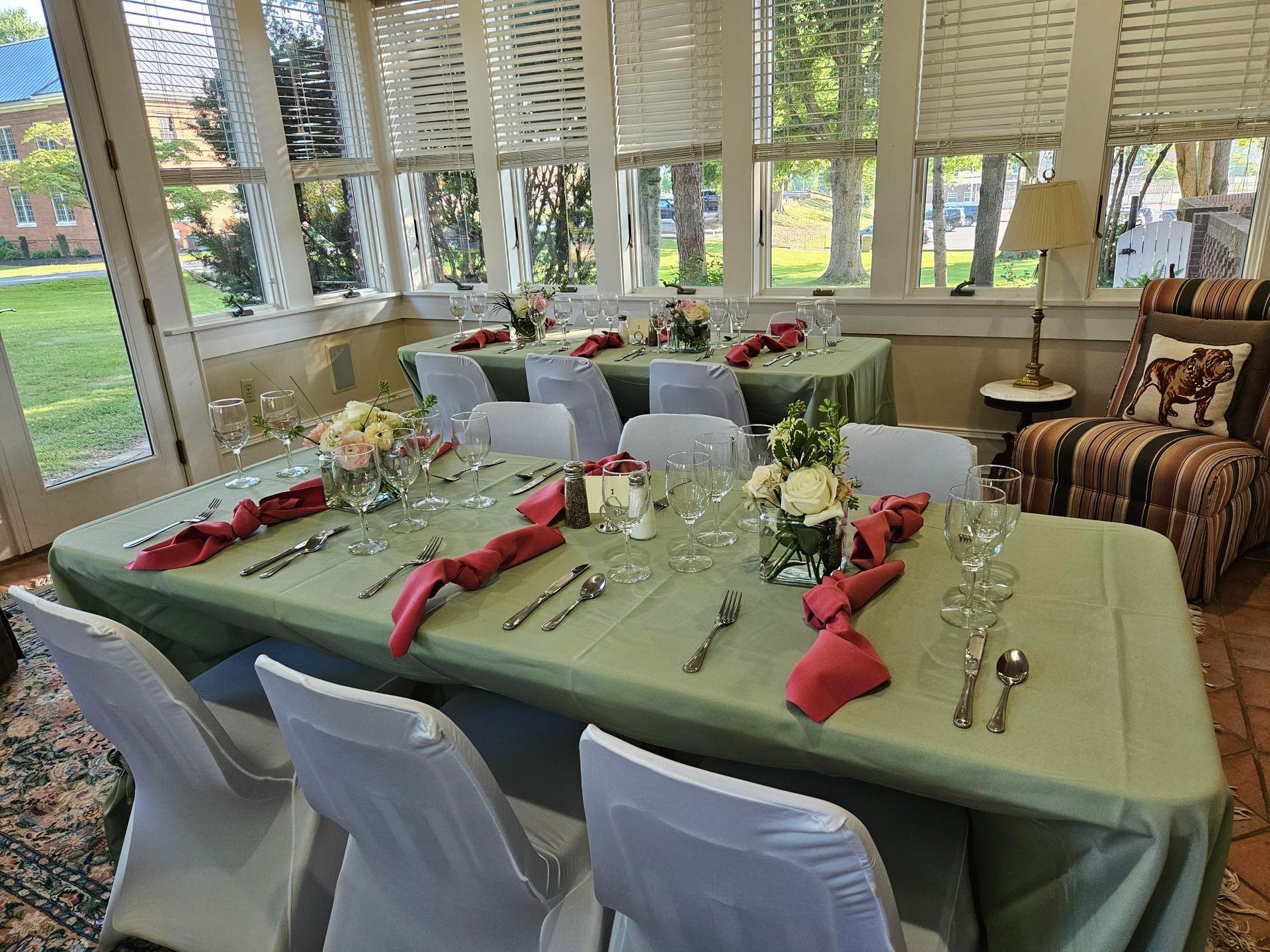 A scene from the wedding reception area inside the sun porch: tables and chairs with place settings including sage green tablecloths, glassware, silverware, rouge cloth napkins, and flower arrangements