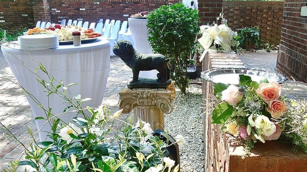 This wedding scene shows a small statue of Mack the Bulldog on a pillar near a table of hors d'oeuvres, floral arrangements on a brick wall, and rows of chairs covered in white cloth in the background.