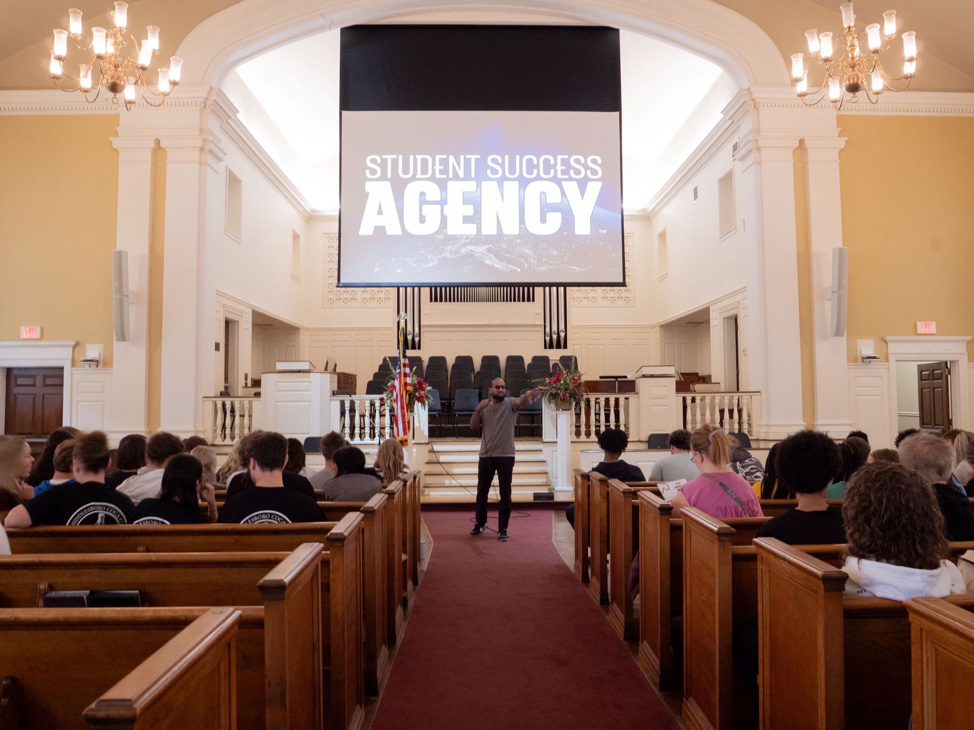 Chris Williams stands in front of the Cowan Chapel stage holding a microphone and pointing out at the audience. The projector screen hanging from the ceiling reads Student Success Agency. Williams is wearing sunglasses and casual attire.