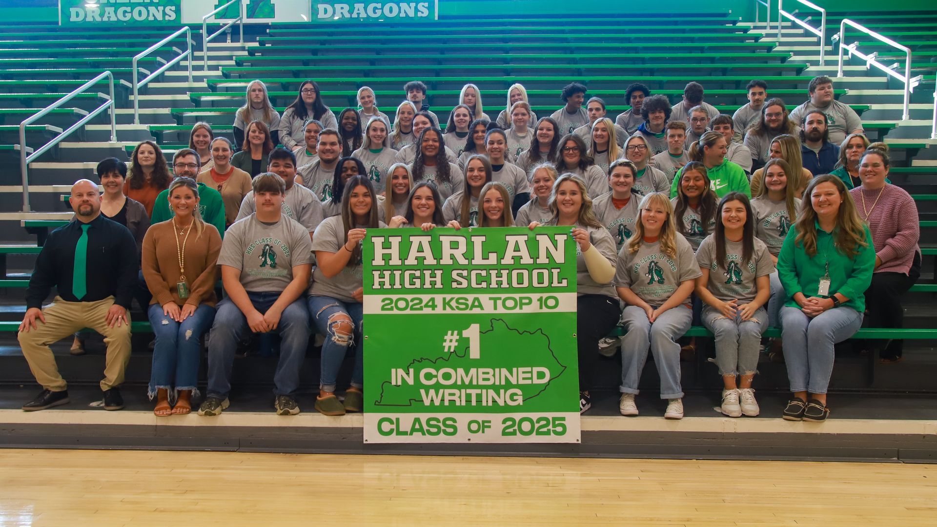 The 11th-grade students who ranked number one on the 2023 KSA on-demand writing exam are wearing shirts that say HHS Class of 2025 and have a green dragon on them. They are all sitting on green bleachers with faculty and staff members holding a sign that says Harlan High School 2024 KSA Top 10 #1 in Combined Writing Class of 2025. Among them are Harlan High School Principal Mike Bolton, sitting on the far left in a black shirt and green tie, and Tyler Saylor, seated behind him, second to the right, wearing a green sweater.